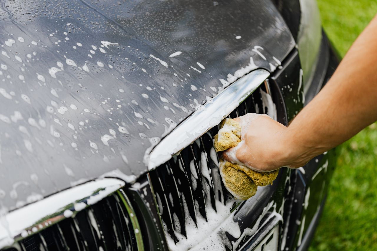 Services-02 Detailed view of a hand washing a car grille with a soapy sponge outdoors.